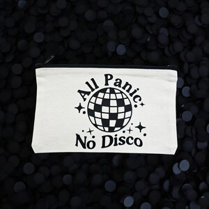 May include: A white canvas zippered pouch with the text "All Panic No Disco" in black. The design includes a checkered globe and star accents. The pouch is rectangular and sits on a dark background.