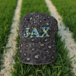 May include: A black baseball cap with a distressed leopard print pattern. The word "JAX" is embroidered on the front in a floral design with yellow and blue accents. The cap is sitting on a green grassy surface.