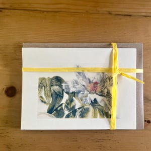 May include: A set of stationery featuring a floral watercolor print, tied with a yellow ribbon. The artwork showcases a detailed depiction of a flower with blue, green, and pink hues, presented on a cream-colored card with a brown envelope.