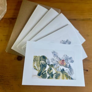 May include: A set of blank greeting cards with floral watercolor art. The cards feature a detailed painting of a white and pink flower with green leaves. The cards are stacked with brown envelopes on a wooden surface.