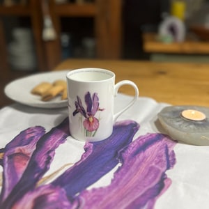 Può includere: Una tazza in ceramica bianca con un motivo floreale di iris viola. La tazza ha un manico curvo e un motivo floreale. La tazza è su una superficie bianca con un grande motivo floreale di iris viola. Un piatto con biscotti è sullo sfondo.
