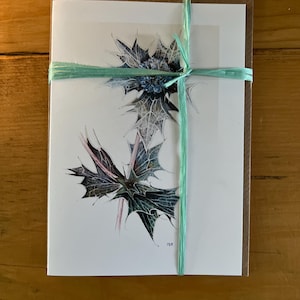 May include: A greeting card with a watercolor illustration of a blue thistle flower. The card is tied with a light green ribbon.
