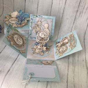 Könnte beinhalten: Eine blaue und weiße Papierbox mit einem Pop-up-Design. Die Box zeigt einen Kelch, ein Kreuz und die Buchstaben "IHS". Die Box ist mit Blumen und Spitze verziert.