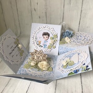 Peut inclure: Une boîte à explosion en carton bleu et blanc avec un garçon en costume tenant un calice. La carte est ornée d'un motif de dentelle et les mots "Première Communion" sur le devant.