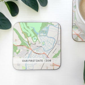 May include: A square coaster with a map of Arundel, England. The map is printed in black and white with a light blue river. The text "OUR FIRST DATE - 2018" is printed in black at the bottom of the coaster.