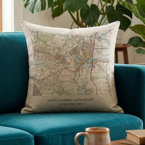 May include: A beige square cushion with a map design, placed on a teal sofa. The map shows roads and buildings in multiple colours. Text at the bottom reads "HAPPY WEDDING ANNIVERSARY 22ND APRIL 2024".