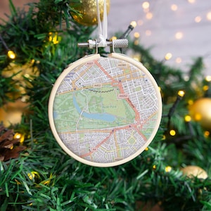 May include: A round, cream-coloured ornament with a map of London, England. The map features Hyde Park, Kensington Gardens, Knightsbridge, Belgravia, and Mayfair. The ornament has a silver hanger.
