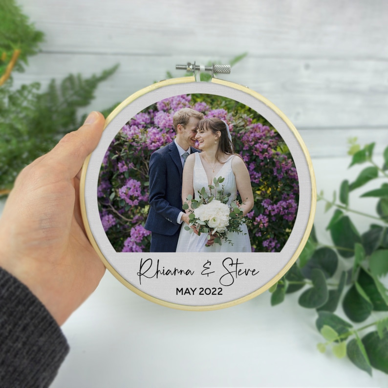 Puede incluir: Un aro de bordado redondo con una foto de una pareja en un entorno de jard&iacute;n. La pareja est&aacute; vestida con atuendo de boda y el texto "Rhianna & Steve MAYO 2022" est&aacute; impreso debajo de la foto.
