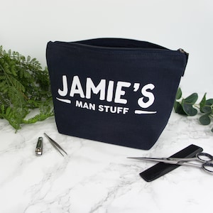 May include: A navy blue zippered toiletry bag with the text "JAMIE'S MAN STUFF" printed in white.