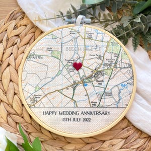 May include: A circular embroidered map in a wooden hoop. The map shows a red heart and the text "HAPPY WEDDING ANNIVERSARY 13TH JULY 2022". The map is beige with brown lines and text.