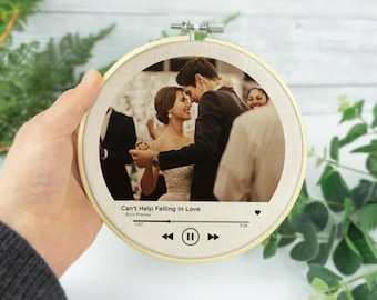 Puede incluir: Un bastidor de bordado redondo con una foto impresa de una pareja bailando. El texto "Can't Help Falling In Love" y un gráfico de reproductor de música están impresos debajo. El bastidor es sostenido por una mano.