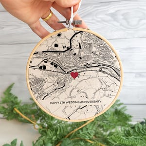 Op de afbeelding: Een ronde, crèmekleurige borduurring met een kaartontwerp en een rood hart. De tekst "HAPPY 4TH WEDDING ANNIVERSARY" is geborduurd aan de onderkant. De ring wordt door een hand vastgehouden.