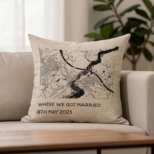 May include: Beige square cushion featuring a black and white map design. The cushion has the text "WHERE WE GOT MARRIED" and "18TH MAY 2023". The cushion is placed on a beige sofa.