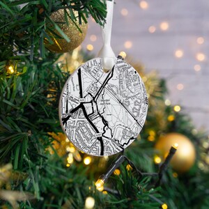 May include: A circular Christmas bauble featuring a black and white map design, suspended by a white ribbon. The map details roads and buildings, with the text "Morden Hall Park". The bauble hangs on a Christmas tree with gold decorations and lights.