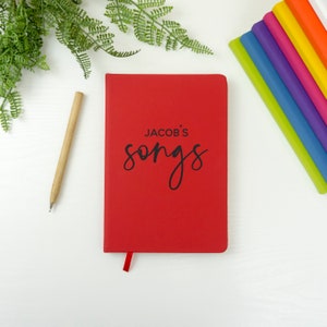 Könnte beinhalten: Rotes Hardcover-Notizbuch mit dem Text "Jacob's songs" in schwarzer Schrift auf der Vorderseite.