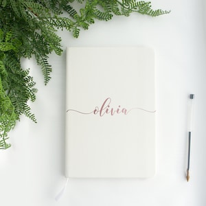 May include: White notebook with a rose gold inscription that reads "olivia" on the front cover. A black pen is lying on a white surface next to the notebook.