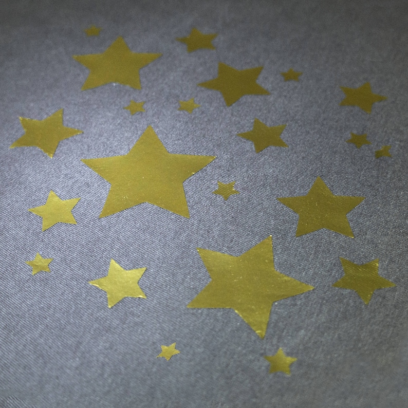May include: A gray fabric with a pattern of gold stars of varying sizes. The stars are scattered across the surface, creating a celestial design. The stars have a metallic sheen, adding a touch of glamour to the fabric.