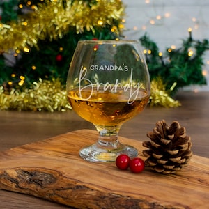 May include: A clear glass brandy snifter with the text "Grandpa's Brandy" etched on the side. The glass is filled with amber-colored liquid and is sitting on a wooden surface with a pine cone and two red berries.