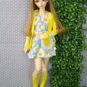 May include: A ball-jointed doll with long blonde hair wearing a light blue dress with a yellow fried egg pattern, a bright yellow cardigan, yellow knee-high socks, white shoes, a white crocheted egg-shaped crossbody bag, and a toast necklace.