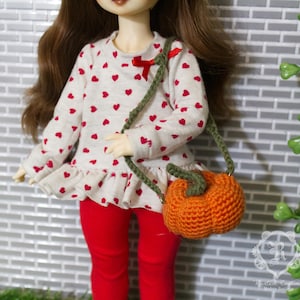 May include: A doll wearing a white long-sleeved shirt with red hearts, red leggings, and a small orange pumpkin-shaped purse with a green strap. The shirt has a red bow at the neckline.