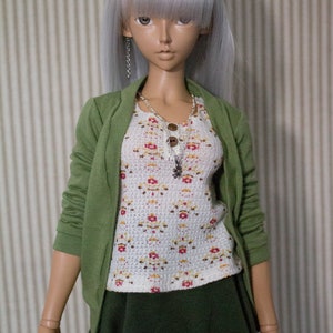 以下が含まれることがあります： 長い銀色の髪をした人形が、緑色のカーディガン、白い花柄のトップス、濃い緑色のスカートを着ています。人形は、チャーム付きの銀色のネックレスをしています。