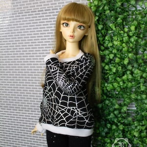 May include: A ball-jointed doll with long blonde hair and bangs, wearing a black long-sleeved shirt featuring a silver spiderweb pattern with white ribbed trim at the neckline and cuffs, paired with solid black leggings.