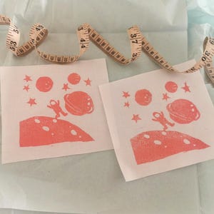 May include: Two white fabric squares with a coral-colored space-themed design. The design includes an astronaut, planets, and stars. A beige measuring tape is draped across the squares.