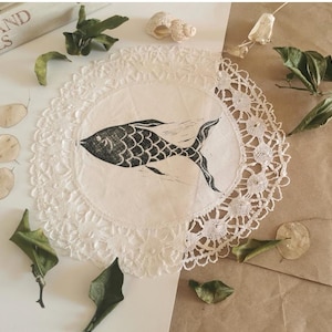 Op de afbeelding: Een witte kanten doily met een zwarte visillustratie. De afbeelding is omgeven door gedroogde bladeren, zaaddozen en een zeeschelp. Op de achtergrond is een boek met de titel "PLANTS AND ANIMALS" te zien.