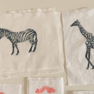 May include: Several white fabric squares with black animal prints. A zebra and a giraffe are depicted. The zebra has black stripes, and the giraffe has a spotted pattern. The fabric has a raw edge.