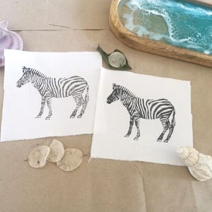 May include: Two white fabric squares with black and white zebra illustrations printed on them.