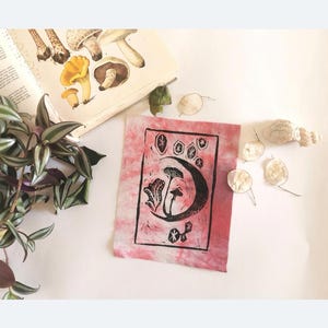 May include: A hand-printed fabric panel with a pink and white tie-dye background. The panel features a black ink block print of mushrooms and a crescent moon.