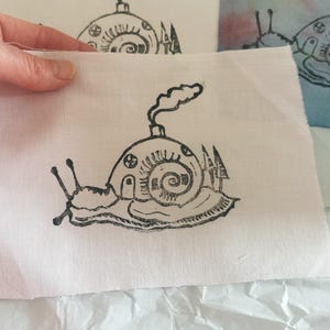 Puede incluir: Grabado en linóleo en blanco y negro de un caracol con una casa en la espalda, con chimenea y árboles. La impresión está sobre una tela blanca. La imagen está sostenida por una mano.