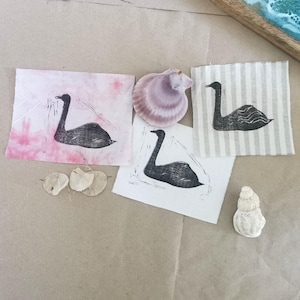 Op de afbeelding: Vier zwarte zwanenstempels op verschillende achtergronden. Twee zijn op wit papier, een op roze papier en een op een gestreepte stof. De stempels zijn allemaal hetzelfde ontwerp, maar de achtergronden zijn verschillend.