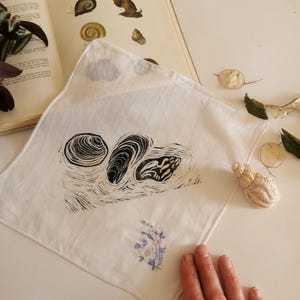 May include: A white cotton handkerchief featuring a black ink print of three seashells. The print is stylised and resembles a woodblock print.