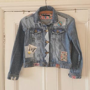 May include: A light blue denim jacket with patches and floral accents. The jacket features a butterfly patch, a bee patch, and floral fabric on the collar and cuffs. The jacket is hanging on a wooden hanger.