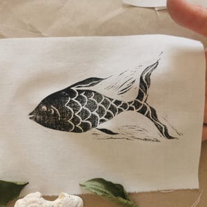 Op de afbeelding: Een zwart-wit linosnede van een vis op een witte stoffen achtergrond.