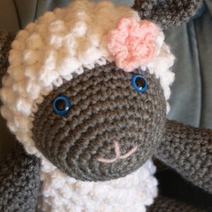 May include: A crocheted stuffed sheep toy with a white fluffy body, gray face, and pink flower on its head. The sheep has blue eyes and a pink nose.