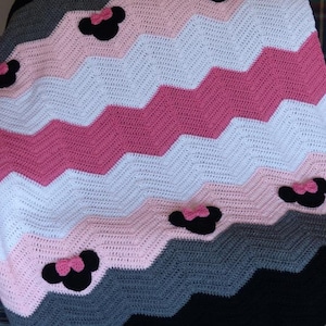 May include: A crocheted baby blanket with a chevron pattern in pink, white, gray, and black. The blanket features several appliqued black and pink Minnie Mouse ears.