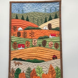 May include: A quilted wall hanging depicting an autumn landscape with rolling hills, houses, trees, and pumpkins. The hills are in shades of orange, yellow, and green. The houses are white with red roofs. The trees are brown with green leaves. The pumpkins are orange. A red tractor is driving on a hill in the background.