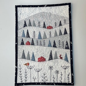 May include: A quilted wall hanging depicting a winter scene with a mountain, trees, and houses. A bird and a deer are also included in the design. The border is dark blue with stars and moons. The color scheme features white, blue, red, and grey.