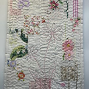 May include: A white fabric wall hanging with a patchwork design featuring pink, green, and white floral patterns. The fabric is quilted and has a delicate, hand-stitched look.
