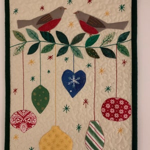 May include: A festive quilted wall hanging with two grey birds and red chests on a green branch. Below, Christmas baubles in red, green, blue, and yellow hang from the branch, with snowflake and star accents.
