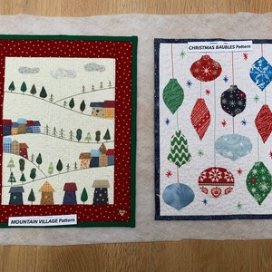 May include: Two fabric art pieces. One features a mountain village scene with trees and houses, labeled "MOUNTAIN VILLAGE PATTERN." The other displays Christmas ornaments, labeled "CHRISTMAS BAUBLES PATTERN." Both are framed with decorative borders.