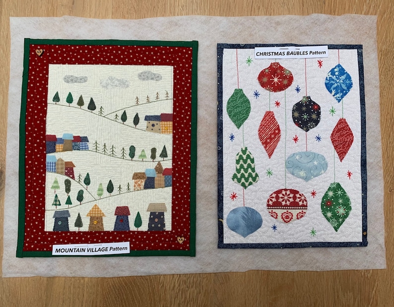 May include: Two fabric art pieces. One features a mountain village scene with trees and houses, labeled "MOUNTAIN VILLAGE PATTERN." The other displays Christmas ornaments, labeled "CHRISTMAS BAUBLES PATTERN." Both are framed with decorative borders.