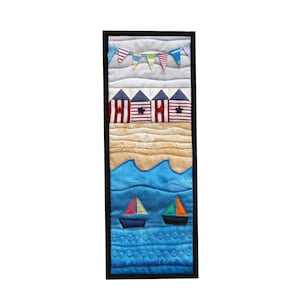 May include: A framed quilted wall hanging depicting a beach scene. The scene includes a blue sky with colourful flags, red and white striped beach huts, a sandy beach, and blue waves with two sailboats.