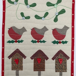 May include: A cream-coloured quilted wall hanging with a red border and white stars. The design features three red and grey birds, three birdhouses with heart-shaped details, and green mistletoe with button embellishments. A festive, handmade item.