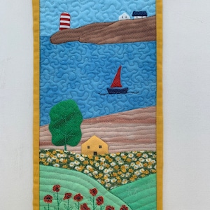 May include: A quilted wall hanging depicting a seascape with a lighthouse, a sailboat, and a small town on a hill. The bottom of the image shows a green field with a yellow house and red poppies.