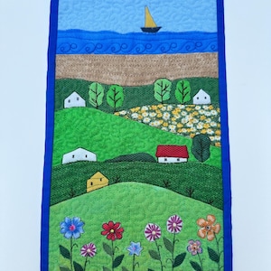 May include: A rectangular fabric wall hanging featuring a quilted landscape scene. The design includes a blue sky with a yellow sailboat, a tan beach, rolling green hills with white houses, a red-roofed cottage, and colourful embroidered flowers.
