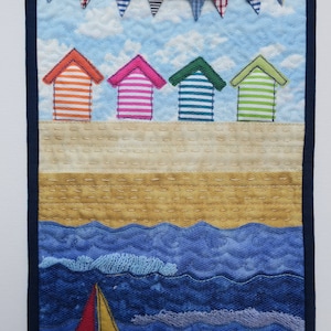 May include: A quilted wall hanging depicting a beach scene with four colourful striped beach huts, a blue sky with white clouds, a string of flags, a sandy beach, and a sailboat in the blue ocean.