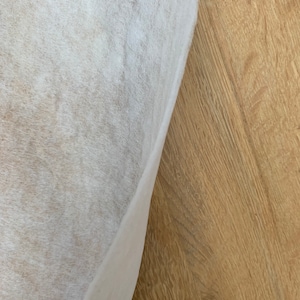 May include: A close-up view of a white, textured fabric partially covering a light brown wooden surface. The fabric has a soft, slightly translucent appearance, with visible fibers. The wood grain is prominent, creating a contrast in texture and color.